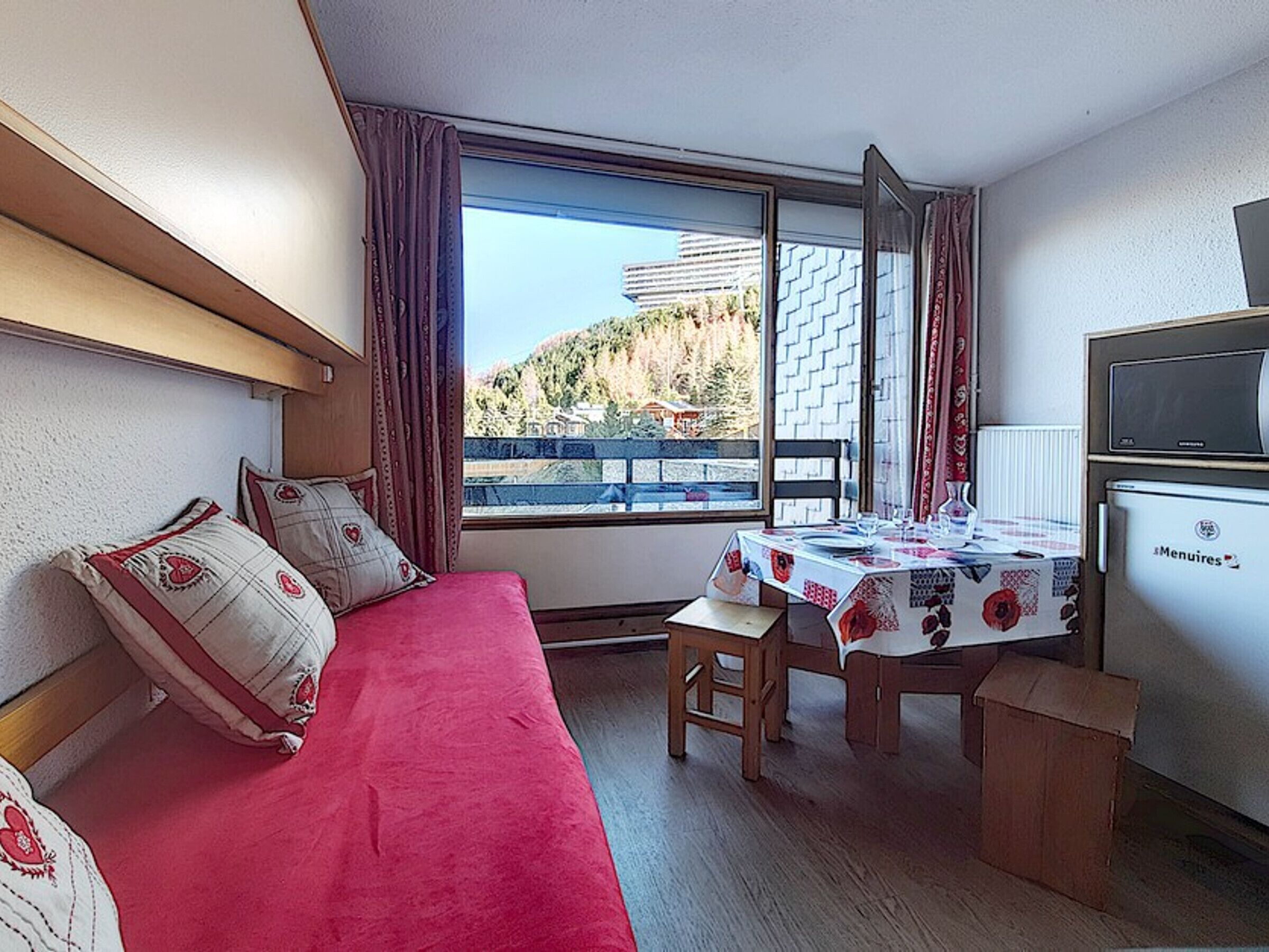 A room with a bed, a table with a tablecloth, a chair, a TV, and a window with a view of buildings and trees.