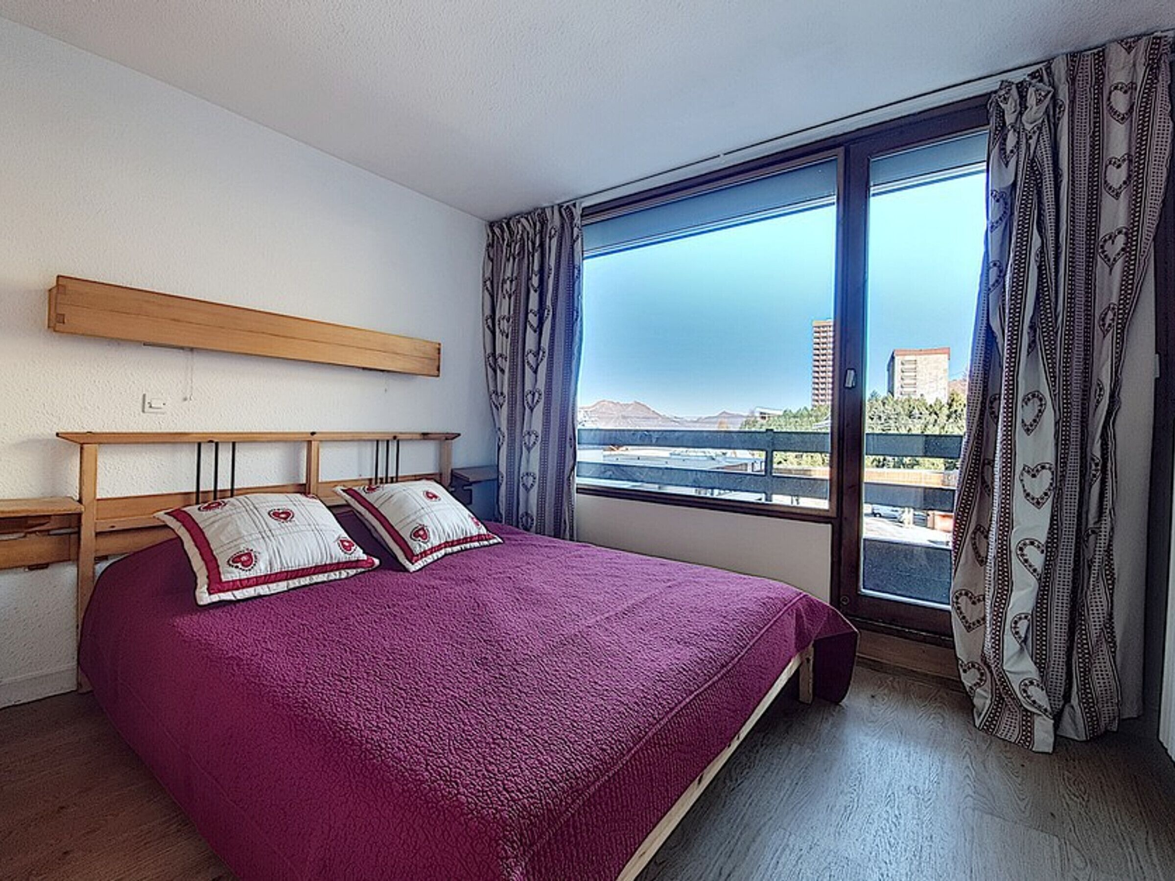 Un dormitorio con una cama grande, cabecera de madera y vistas a un paisaje urbano a través de un amplio ventanal.