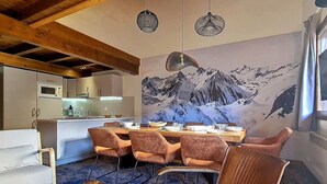 Une cuisine moderne avec des meubles blancs, un coin repas doté d’une table en bois et de chaises en velours, et une fresque représentant une montagne sur le mur.