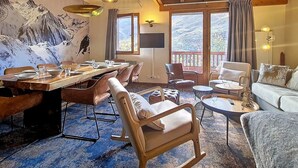 Un coin repas avec une table en bois dressée pour quatre personnes, entourée de chaises rembourrées. On y trouve une grande fenêtre avec des rideaux, un canapé avec des coussins et une petite table ronde avec un verre.