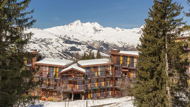 Un chalet à étages, doté d’une façade en bois et de balcons, entouré de montagnes enneigées et d’arbres à feuilles persistantes.