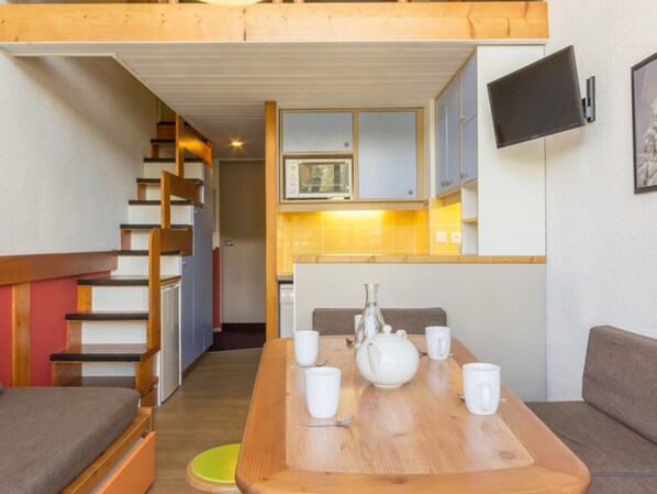 Un espace de vie compact comprenant une table à manger pour quatre personnes, un canapé, un escalier et un coin cuisine avec un téléviseur fixé au mur.