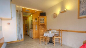 Une pièce chaleureuse avec un plancher en bois, une table à manger avec des chaises, un lit et un coin cuisine visible à travers une porte ouverte.