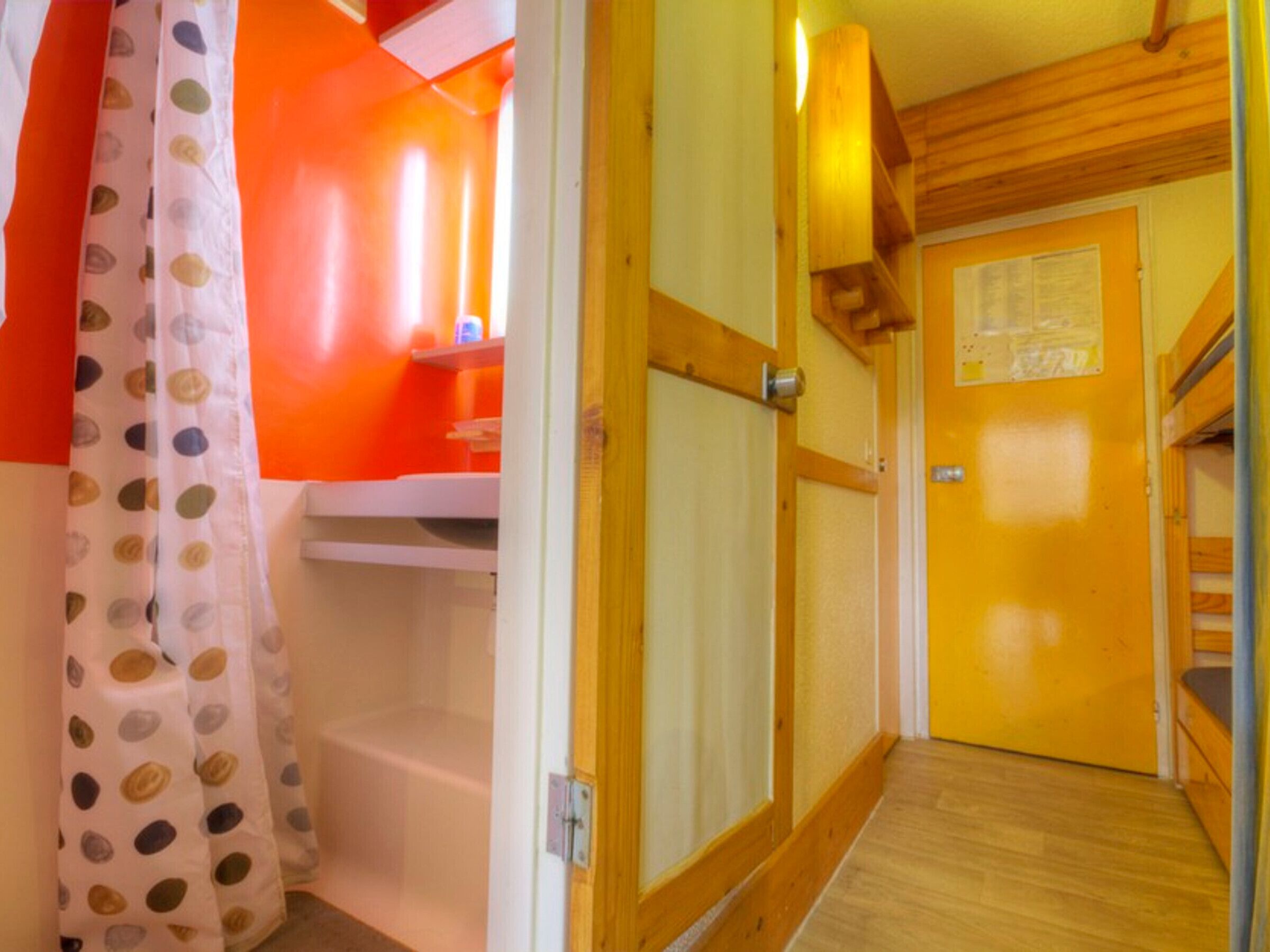 Un couloir étroit avec une porte en bois, un mur blanc orné d’un tableau d’affichage, et une salle de bain avec un mur orange et un rideau de douche à motifs à pois.