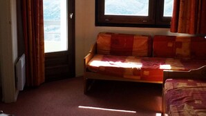 Et værelse med en sofa, et fjernsyn ophængt på væggen og et vindue med gardiner.