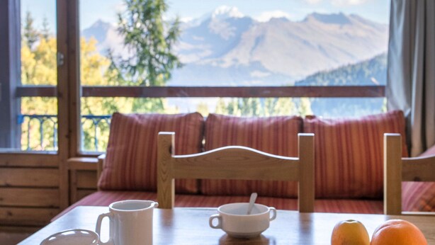Une table en bois sur laquelle se trouvent une tasse blanche, une autre tasse blanche avec une cuillère, et deux oranges. Une chaise en bois est placée derrière la table. La pièce offre une vue sur les montagnes grâce à de grandes fenêtres.