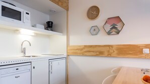 Una cocina compacta con microondas, cafetera y lavavajillas. Cuenta con un revestimiento de pared de madera y una pequeña mesa con una cesta de fruta.