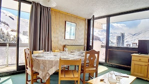Un comedor con una mesa dispuesta para cuatro, sillas de madera, una mesita con un bol y copas de vino, y grandes ventanales con vistas a una montaña nevada.