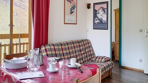 Um quarto aconchegante com um sofá xadrez, uma mesa de jantar posta com pratos, xícaras e um jarro, e um pôster de uma paisagem de montanhas.