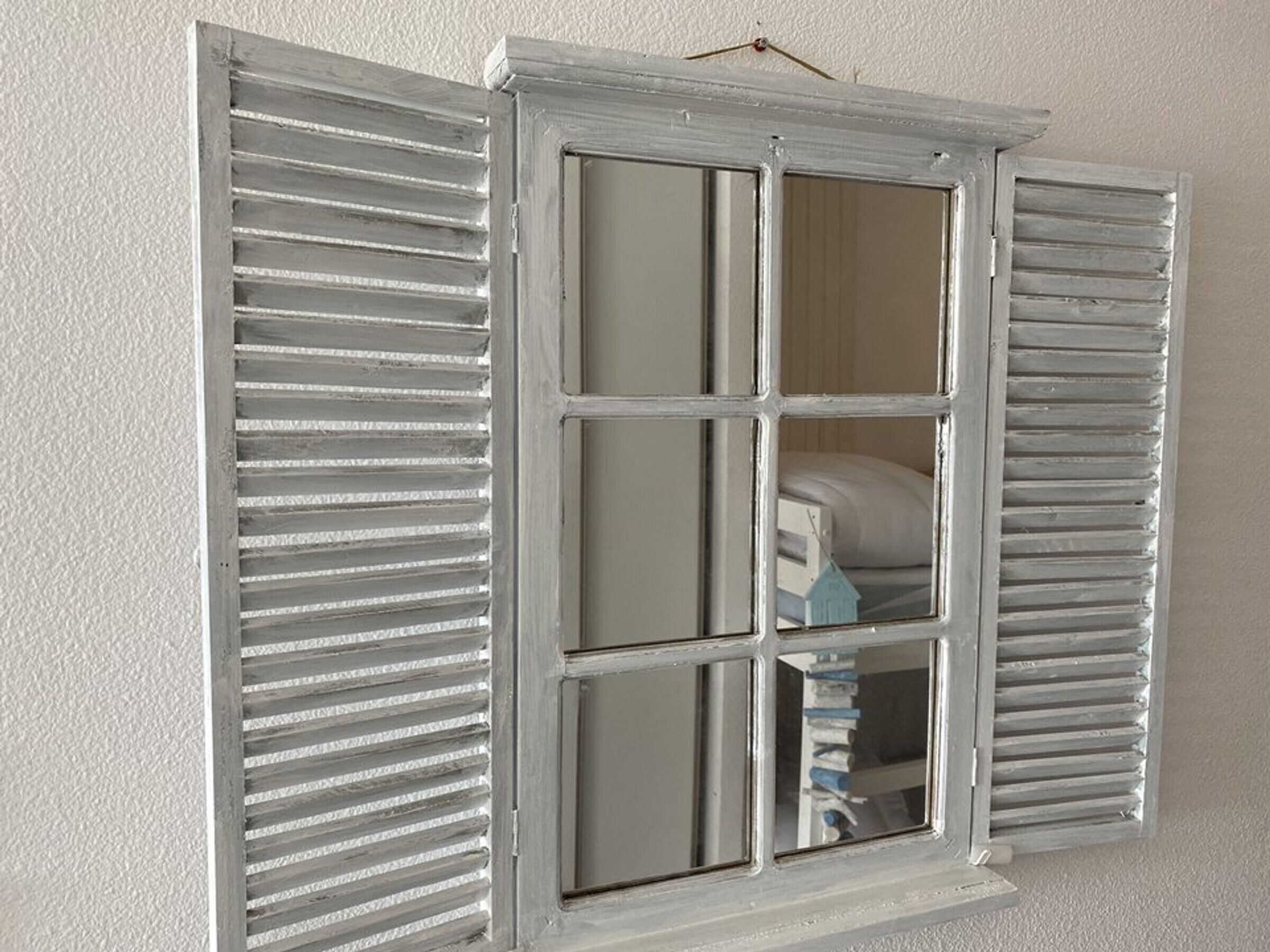 Une fenêtre avec des volets blancs, divisée en quatre carreaux, dans laquelle on aperçoit un lit et un objet bleu à l’intérieur.