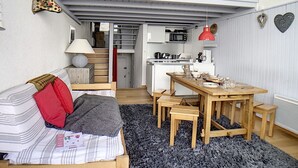 A compact, multi-level room with a loft bed, a dining area with a wooden table and benches, a kitchen area with modern appliances, and a cozy seating area with a sofa and pillows.