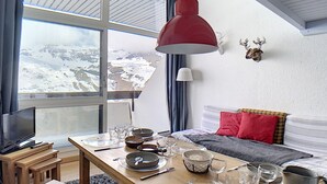 A cozy room with a wooden dining table set for four, a bed with red and gray bedding, a mounted deer head on the wall, and a large window offering a snowy mountain view.