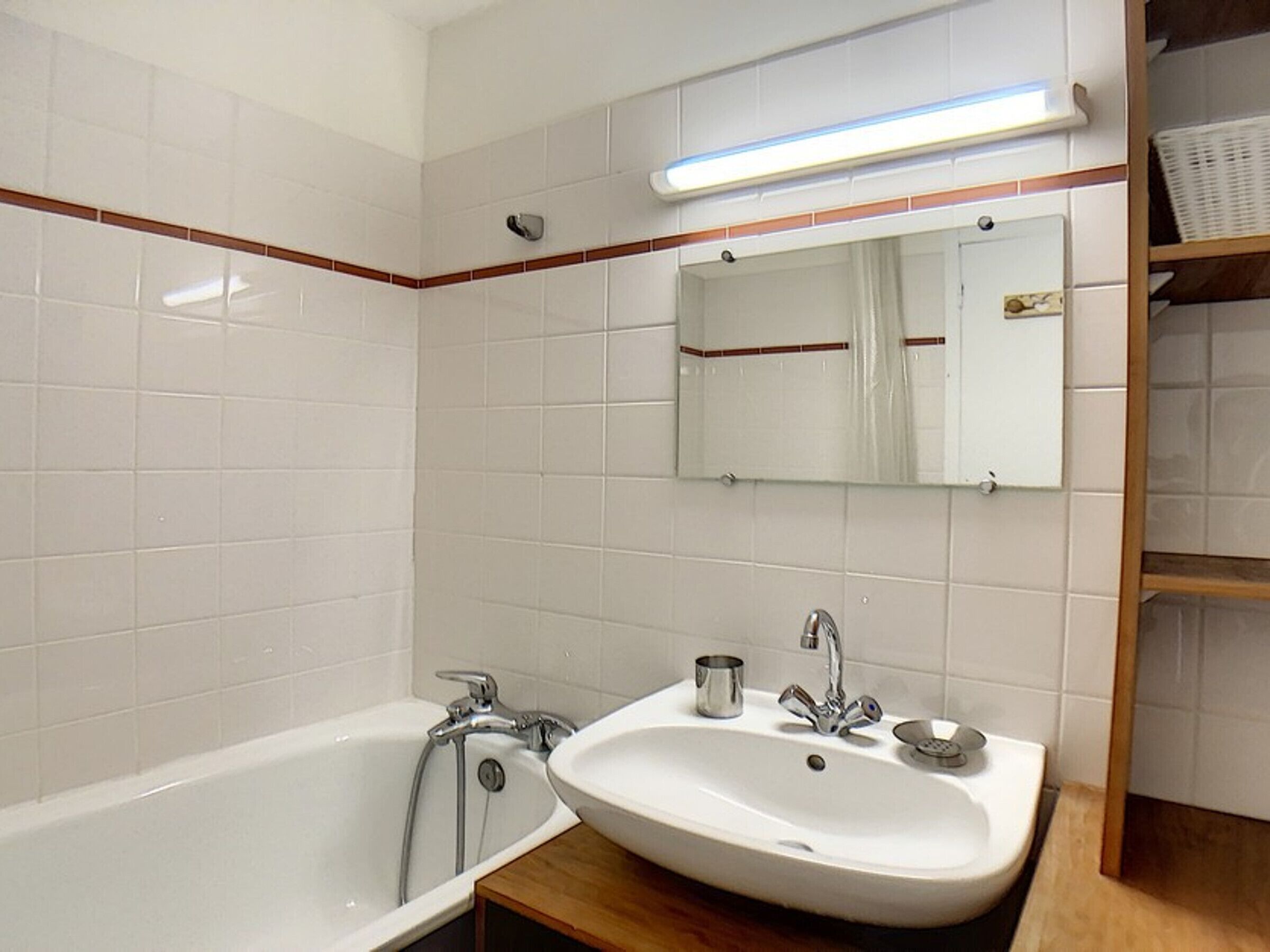 Ein Badezimmer mit weißen Fliesen, einer Badewanne, einem Waschbecken, einem Spiegel und einem Regal mit einem Korb.