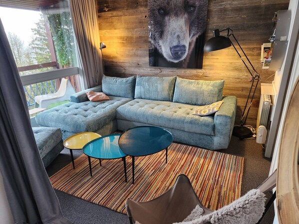 Ein gemütliches Wohnzimmer mit einem Ecksofa, zwei kleinen runden Tischen und einem großen Bärenbild an der Wand.