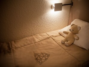 A single bed with a pillow and a teddy bear, a wall-mounted light, and a geometric design on the bedspread.