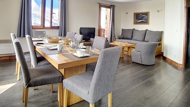 A modern living room with a dining area, featuring a wooden dining table, grey upholstered chairs, a grey sofa, and a television.