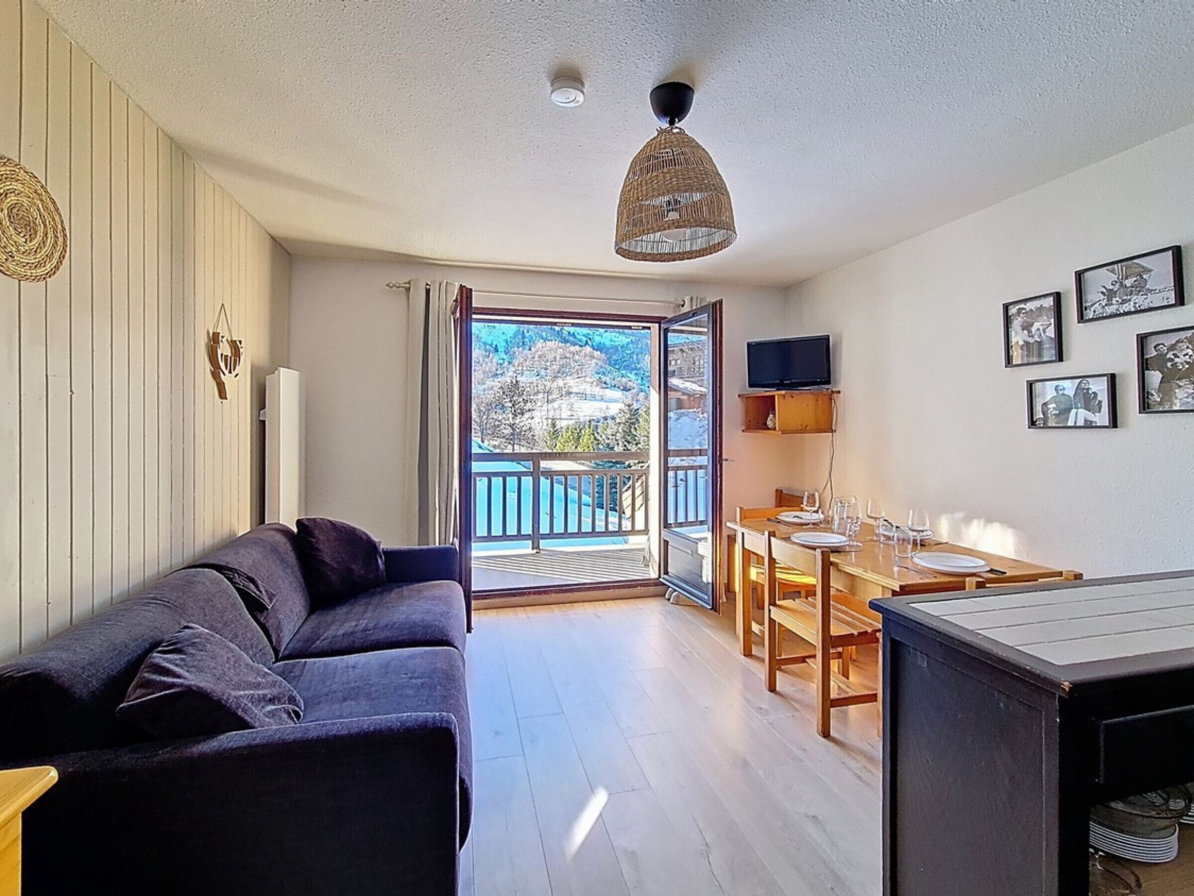 A modern living room with a dark sofa, a dining area with wooden furniture, and a balcony with a mountain view.