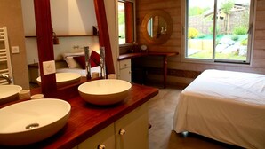 Ein Schlafzimmer mit einem großen Bett, zwei Waschbecken, einem Waschtisch mit Spiegel und einem Fenster mit Blick auf Grünflächen.