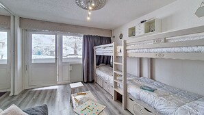 A room with bunk beds, a small table with a board game, a radiator, and a window with a view of a snowy landscape. - Central apartment with balcony for 6 people (Les Menuires)