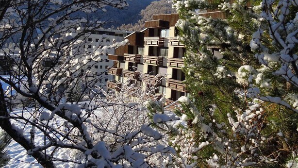 Ein mehrstöckiges Gebäude mit Balkonen, umgeben von schneebedeckten Bäumen und Bergen im Hintergrund.