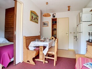 Quarto compacto com cama de madeira, pequena mesa de jantar com cadeiras, área de cozinha com geladeira e porta que dá para outro cômodo.