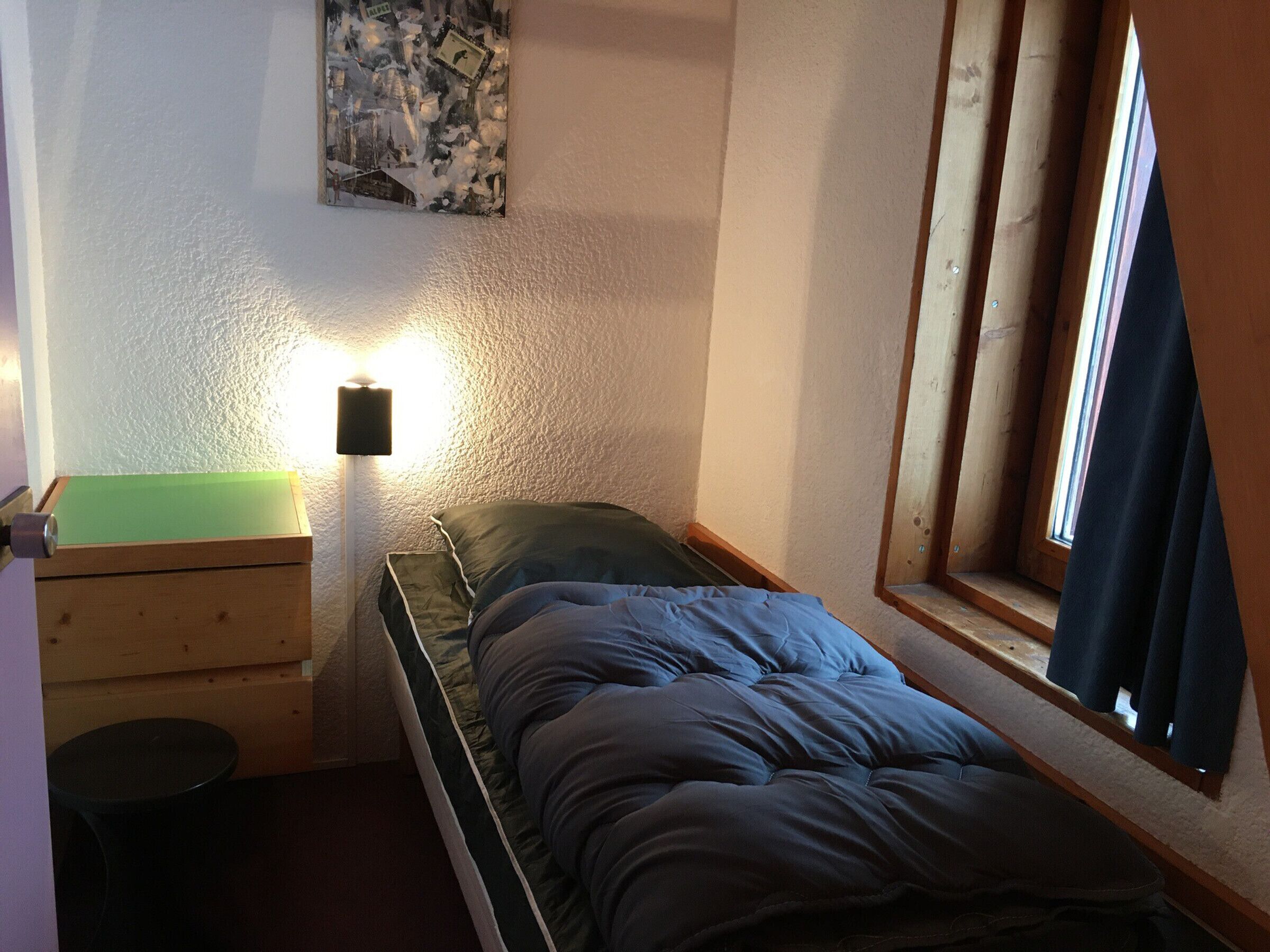 Una stanza piccola con un letto singolo, una testiera in legno, un comodino, una lampada e una finestra con tende.