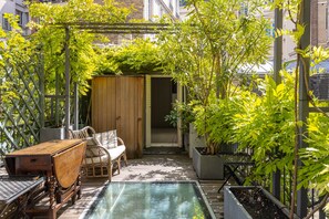 A sunlit patio with a wooden table and chairs, a small pool, and lush greenery.