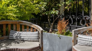 A wooden bench with a cushion, a planter with plants, and a metal fence in a courtyard. - The Arbour, by Veeve (Paris)