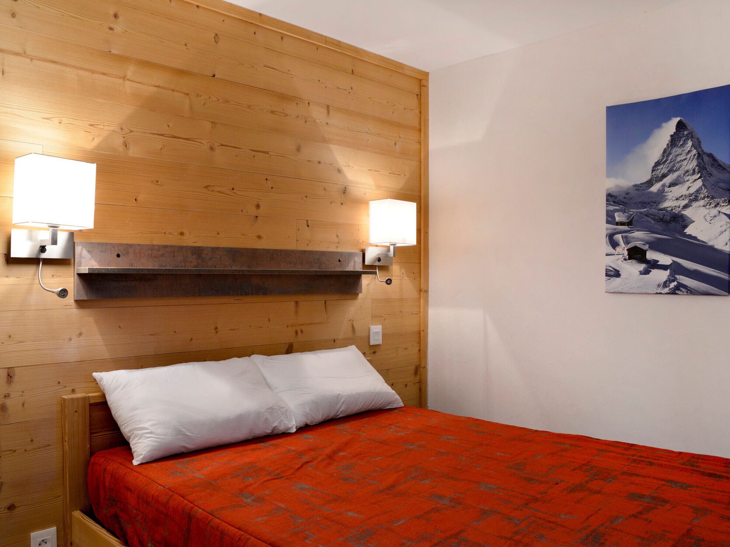 A hotel room with wooden paneling, a bed with white and orange bedding, two wall-mounted lights, and a mountain landscape painting.