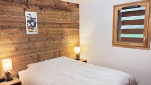 A bedroom with a wooden headboard, a bed with white bedding, two bedside lamps, and a window with blinds. - Les Gets: 2-bed apartment with terrace and WIFI, near center and slopes (Les Gets)