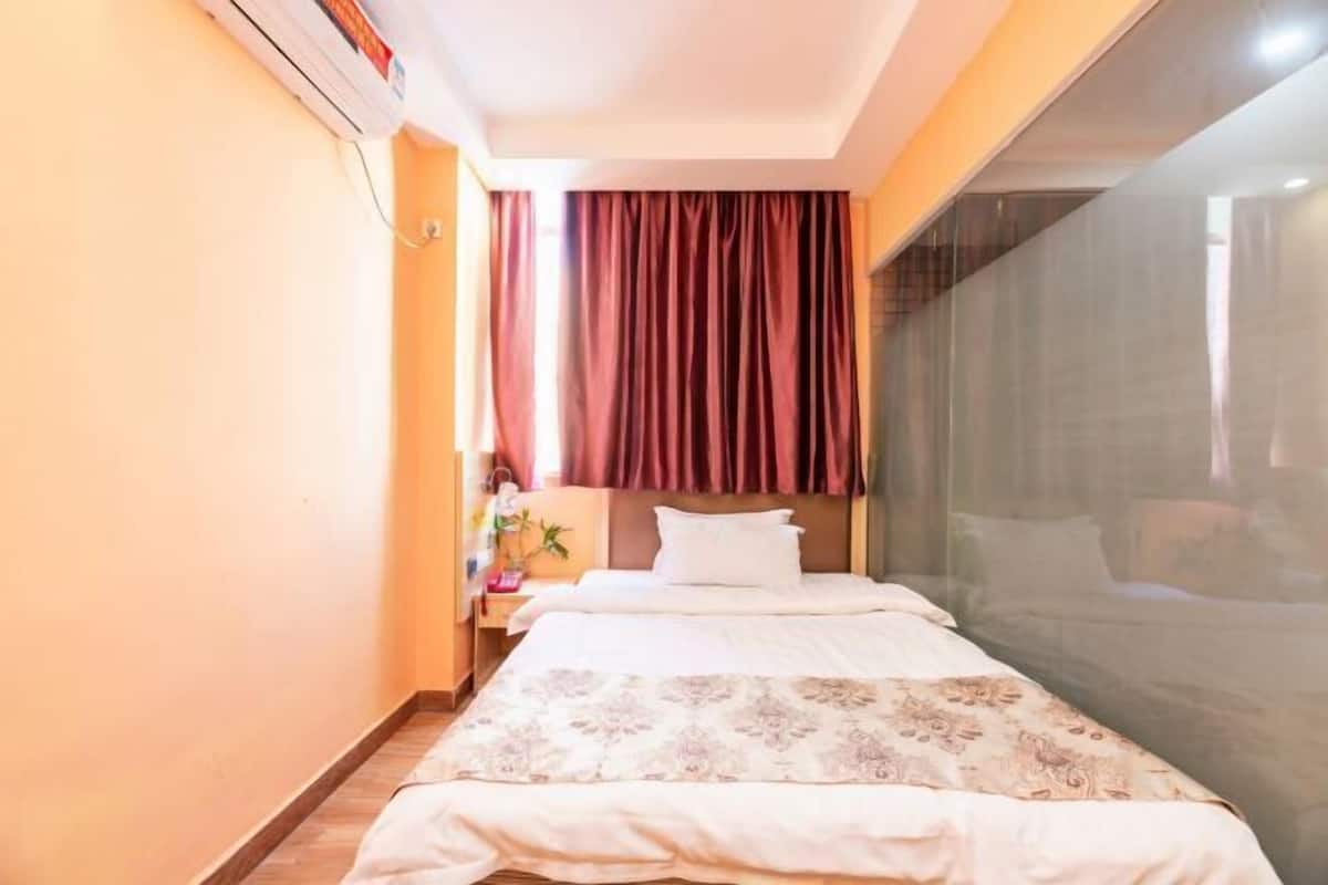 A hotel room with a bed, a nightstand, a window with curtains, and a wall-mounted air conditioner.