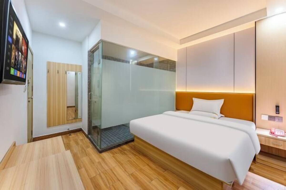 A modern hotel room with a bed, a glass shower enclosure, a wooden floor, and a wall-mounted TV.