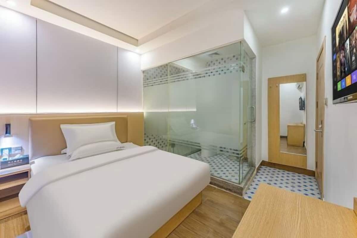 A hotel room with a bed, a glass-enclosed shower, a wooden bedside table, and a wall-mounted TV.