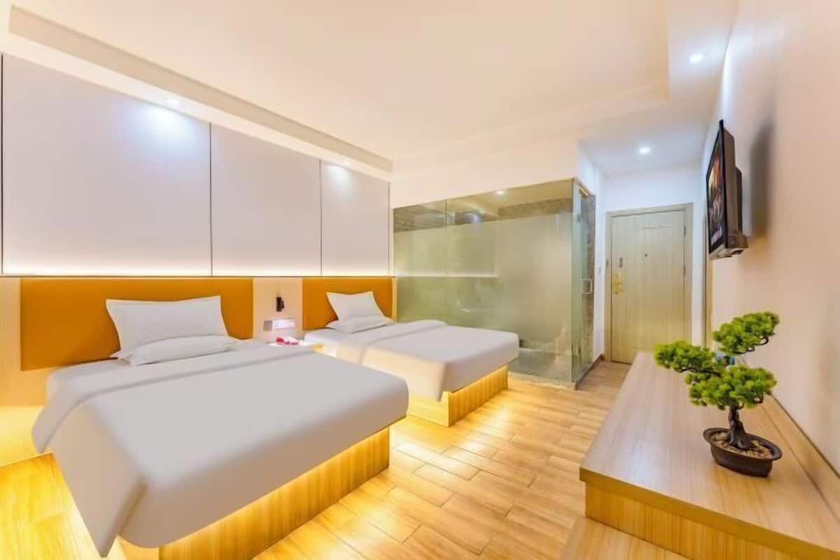 A modern hotel room with two beds, a wooden floor, a small table with a plant, and a TV mounted on the wall.