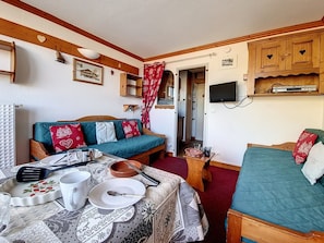 Quarto aconchegante com cama de madeira, sofá com almofadas estampadas de coração, mesa de jantar com pratos e talheres, e televisão na parede.