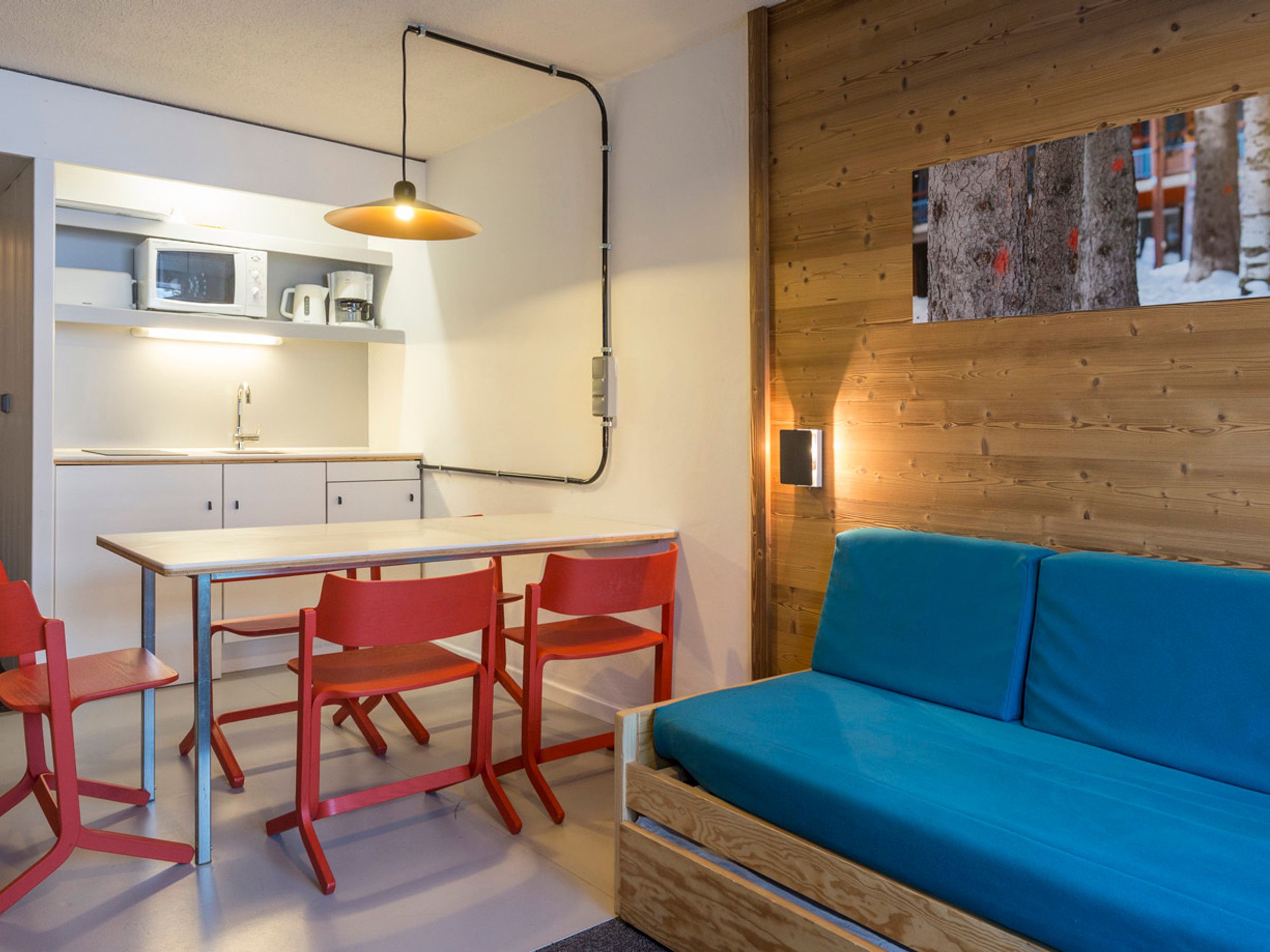 A compact living space with a kitchenette, a dining table with red chairs, and a blue sofa against a wooden wall.