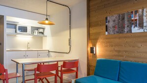 Een ruimtelijk ingerichte leefruimte met kitchenette, een eettafel met rode stoelen en een blauwe bank tegen een houten wand.