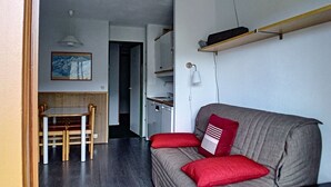 A compact living space with a sofa, red cushions, a dining table with chairs, and a kitchen area with a microwave and a hanging light fixture.