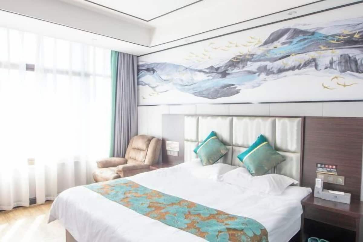 A hotel room with a large bed, a chair, a painting on the wall, and a window with curtains.