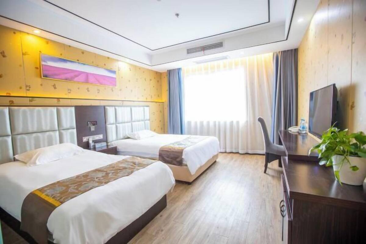 A hotel room with two beds, a desk, a chair, a TV, and a large window with curtains.