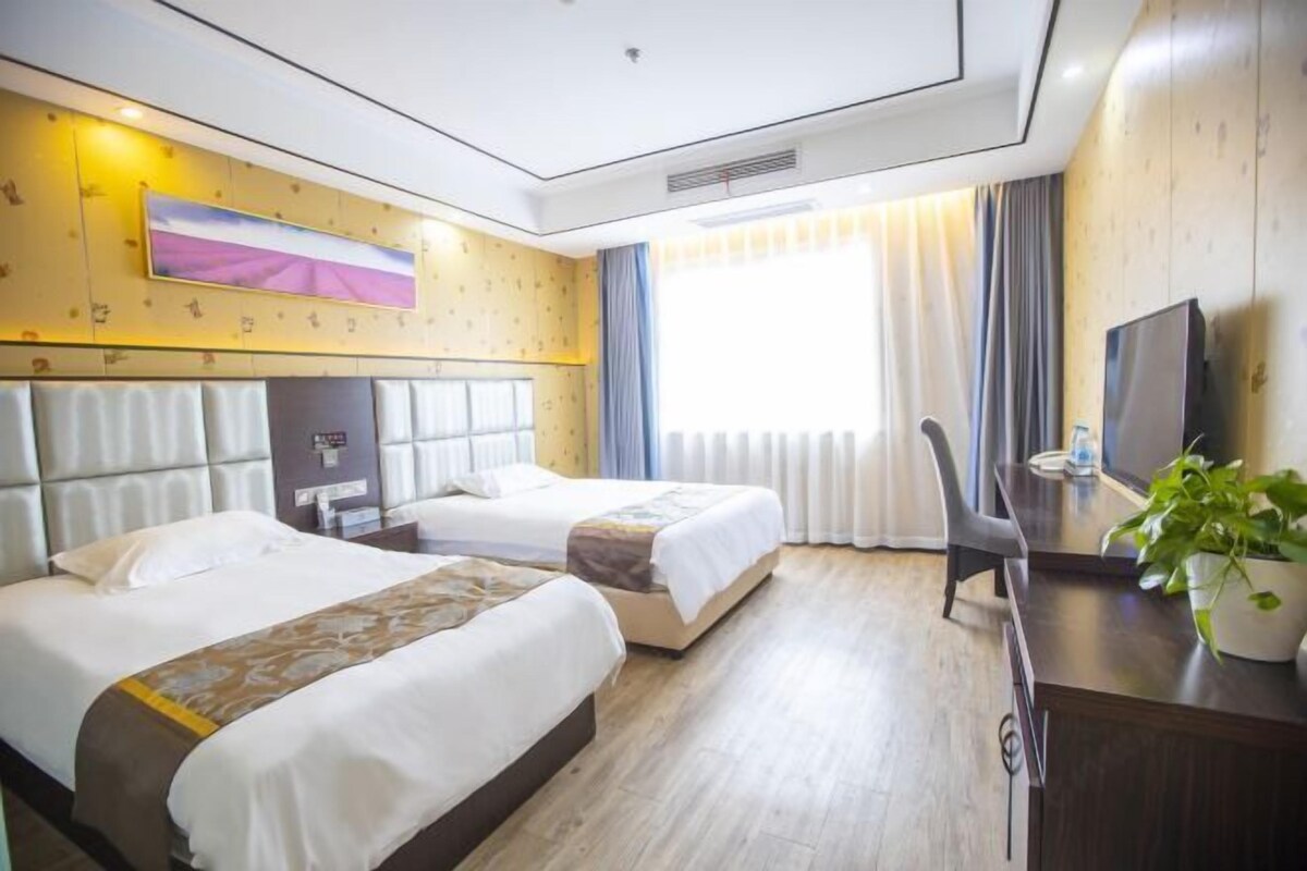 A hotel room with two beds, a desk, a chair, a TV, and a large window with curtains.