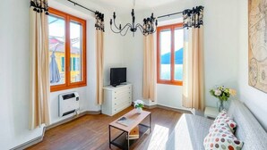 Une pièce avec un plancher en bois, une commode blanche, un téléviseur, une petite table, un canapé avec des coussins à pois et de grandes fenêtres avec des rideaux.
