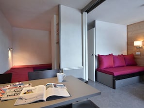 Quarto de hotel compacto com uma cama, uma mesinha com uma revista e uma parede branca com luminária.