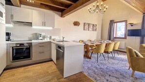 A modern kitchen with white cabinets, a built-in oven, and a dishwasher. There is a dining area with a table and chairs, and a chandelier above.