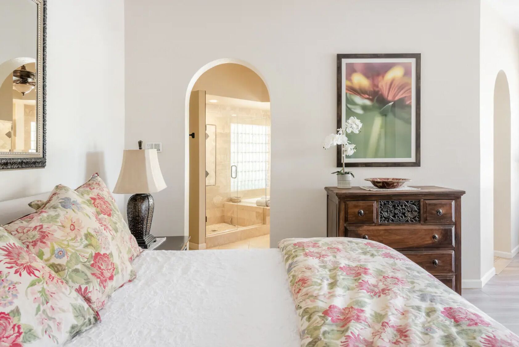 A hotel room with a bed, a dresser, a lamp, a framed picture, and a shower area visible through an archway.