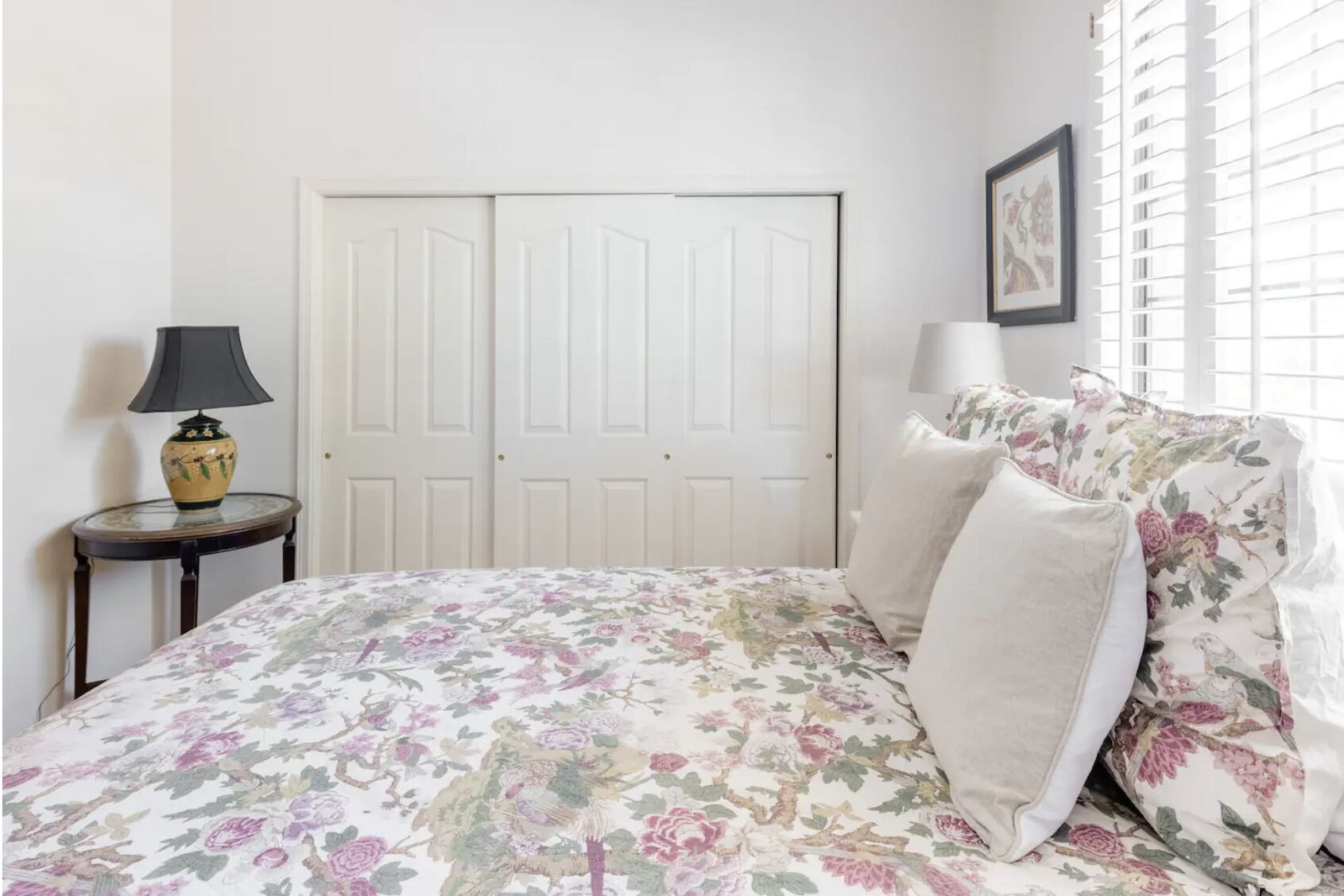 Une chambre à coucher avec une couvre-lit à motifs floraux, deux lampes de chevet, une table ronde avec une lampe et une porte à deux battants.