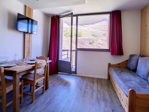 Una habitación con una mesa de comedor de madera dispuesta para dos personas, un sofá, un televisor montado en la pared y un ventanal con cortinas.