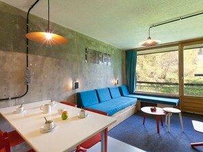 A modern living room with a blue sofa, a dining table with cups and a fruit, and a large window with a view of trees.