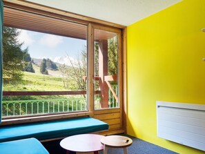 Une pièce avec un banc en bois, une petite table ronde et une vue sur un paysage verdoyant, visible à travers une grande fenêtre.