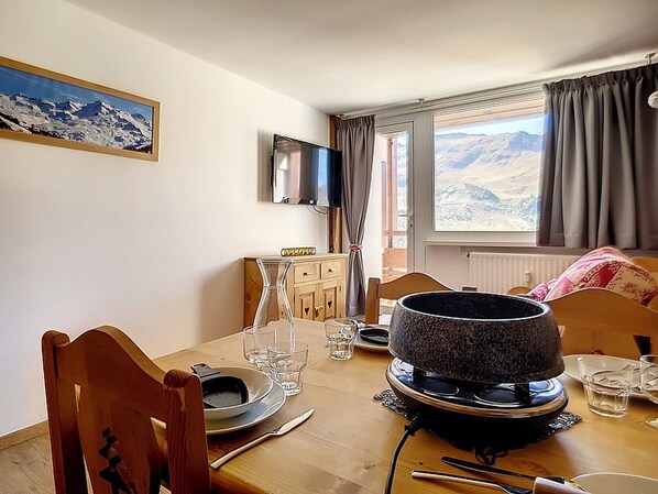 A dining area with a table set for two, a television mounted on the wall, and a window with a view of mountains.